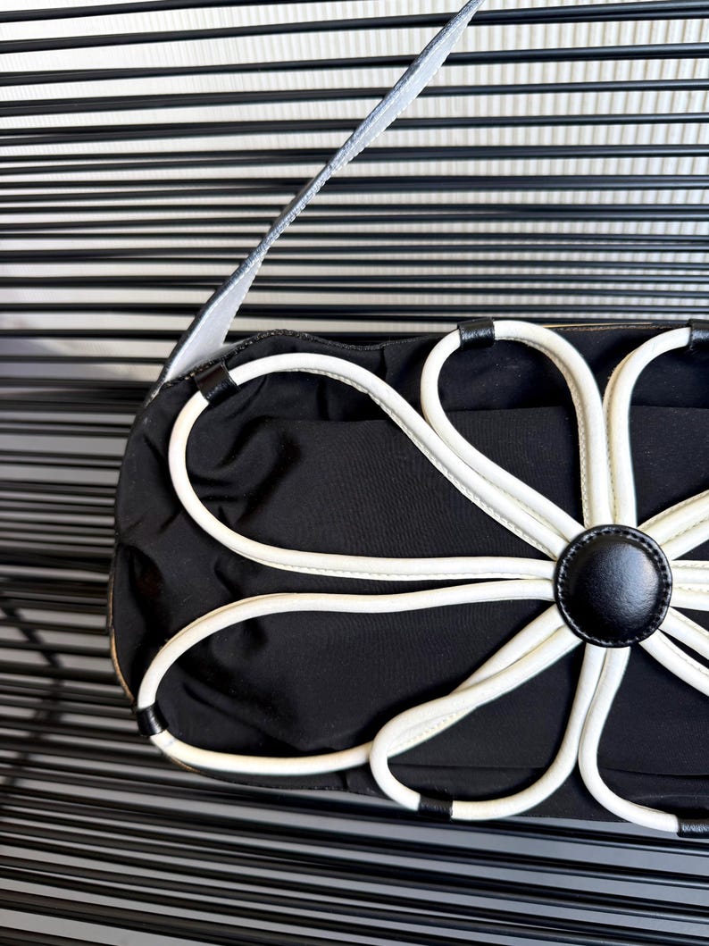 Puede incluir: Un bolso de mano negro con una prominente aplicaci&oacute;n floral blanca en la parte frontal. El dise&ntilde;o de flor se compone de p&eacute;talos de ribete blanco que irradian desde un bot&oacute;n circular negro central. El bolso tiene una correa negra y cuerpo liso.