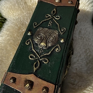 May include: A dark green, rectangular book with a bear design on the cover. The cover has copper-colored metal accents and decorative rivets. The book is resting on a fluffy, cream-colored surface.