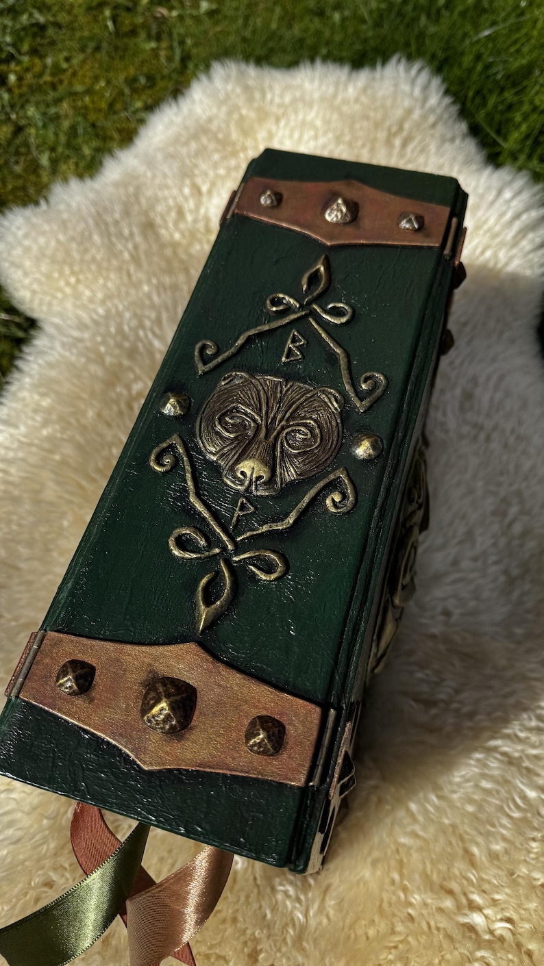 May include: A dark green, rectangular book with a bear design on the cover. The cover has copper-colored metal accents and decorative rivets. The book is resting on a fluffy, cream-colored surface.