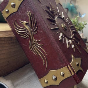 May include: A burgundy-colored book with gold-colored metal accents. The cover features a phoenix design and a sunburst emblem. The edges are trimmed with gold metal and studded with small, gold-colored rivets. A cream-colored ribbon serves as a bookmark.