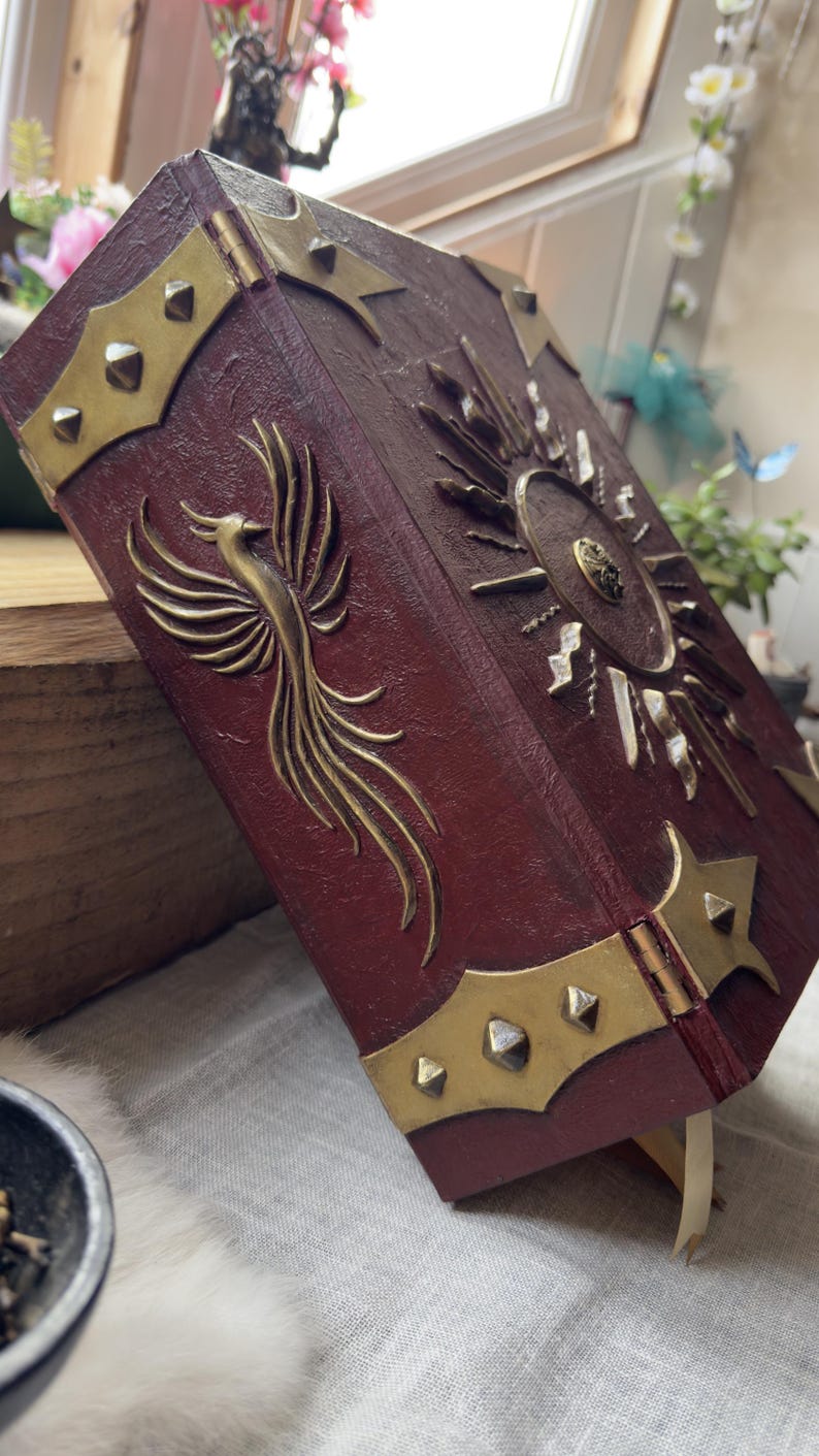 May include: A burgundy-colored book with gold-colored metal accents. The cover features a phoenix design and a sunburst emblem. The edges are trimmed with gold metal and studded with small, gold-colored rivets. A cream-colored ribbon serves as a bookmark.