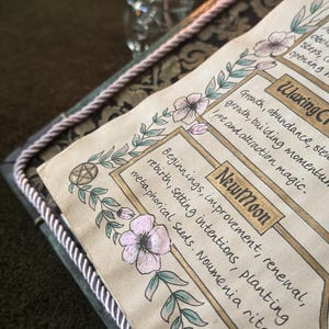 K&ouml;nnte beinhalten: Eine Seite aus einem Grimoire mit handgezeichneten Blumenr&auml;ndern und Text in Goldtinte. Der Text lautet New Moon, Beginnings, improvement, renewal, rebirth, setting intentions, planting metaphorical seeds, Noumenia rite, Waxing Crescent, Growth, abundance, steady growth, building momentum, love and attraction magic.