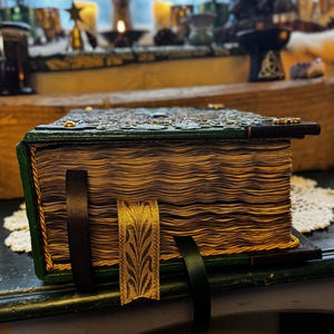 Może przedstawiać: Stylowa, sk&oacute;rzana książka w stylu vintage z zieloną okładką i złotymi akcentami. Książka ma zapięcie na zatrzask i kilka wstążkowych zakładek. Strony są postarzałe, a książka spoczywa na ciemnej powierzchni.