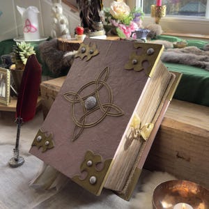 May include: A large, open book with a textured, mauve cover and ornate brass corner pieces. A Celtic knot design adorns the front. The book is propped open on a wooden surface, with a red feather quill nearby.