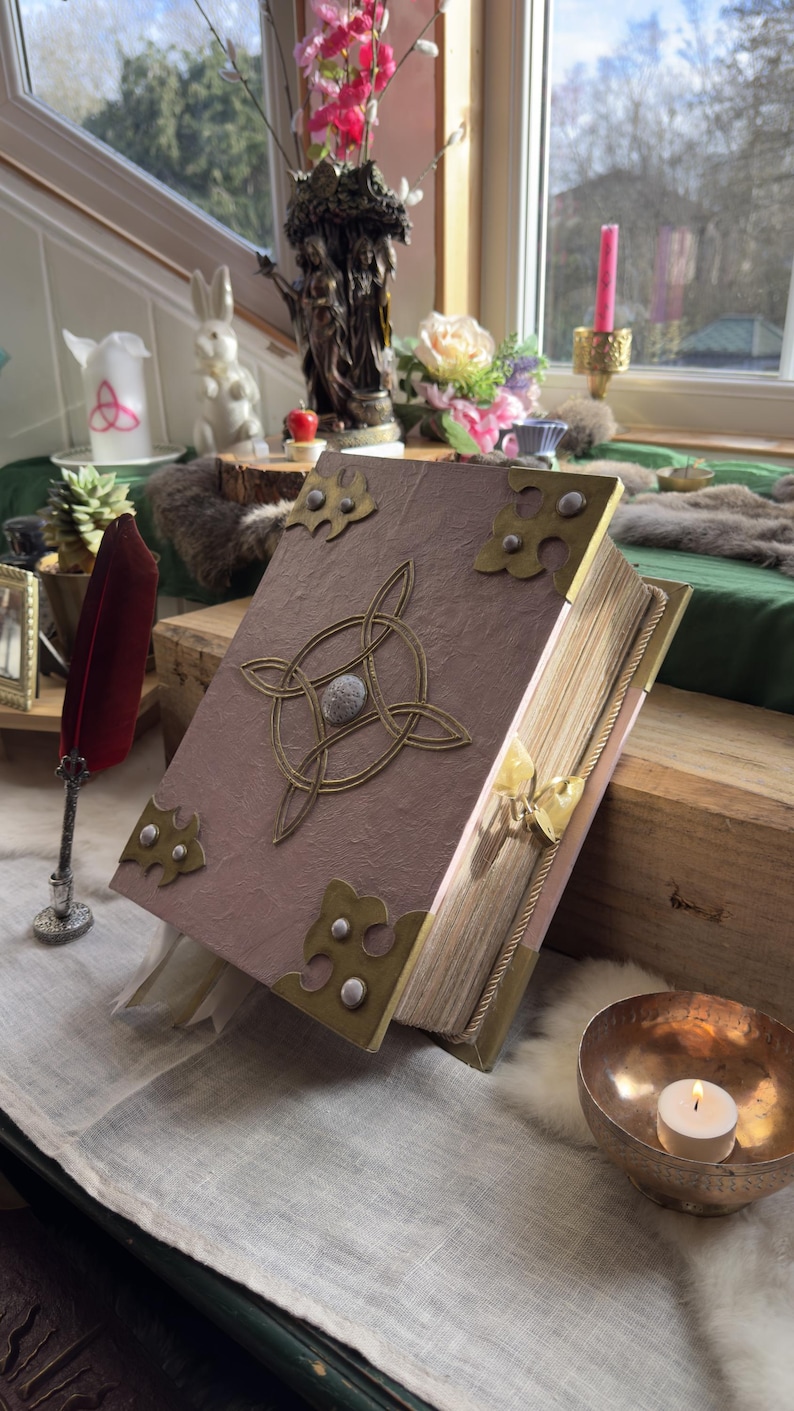 May include: A large, open book with a textured, mauve cover and ornate brass corner pieces. A Celtic knot design adorns the front. The book is propped open on a wooden surface, with a red feather quill nearby.