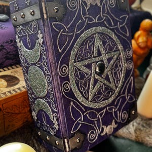 May include: A purple, decorative book with silver Celtic knot and pentagram designs. The book has metal corner protectors and hinges. A silver crescent moon design is on the side. A lit candle and other decorative items are in the background.