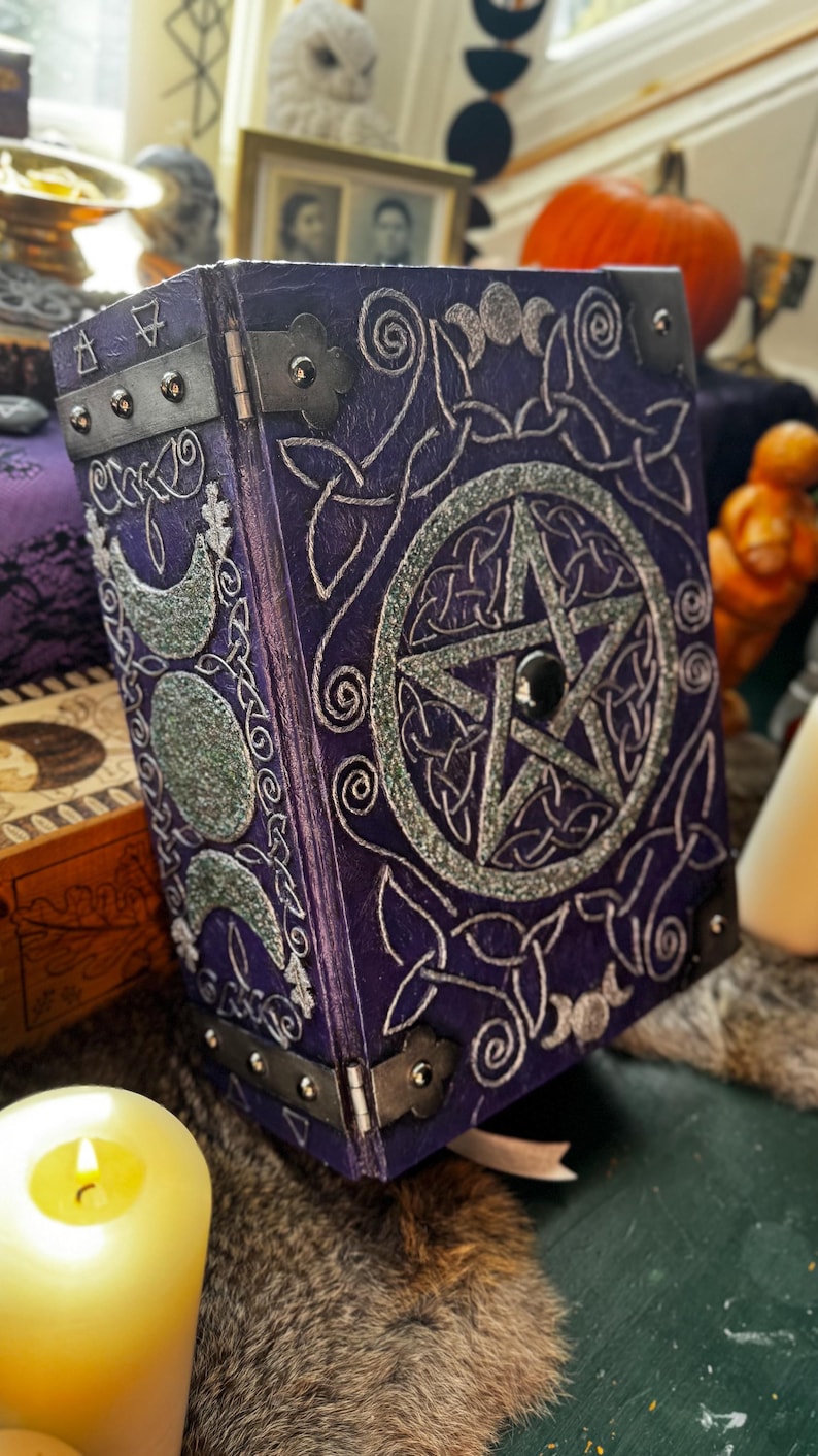 May include: A purple, decorative book with silver Celtic knot and pentagram designs. The book has metal corner protectors and hinges. A silver crescent moon design is on the side. A lit candle and other decorative items are in the background.