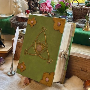 May include: A green book with a gold Celtic knot design and pink accents. The book has gold corner pieces and a ribbon closure. The pages are edged with lace. The book is propped open on a table with a white feather quill and other decorative items.