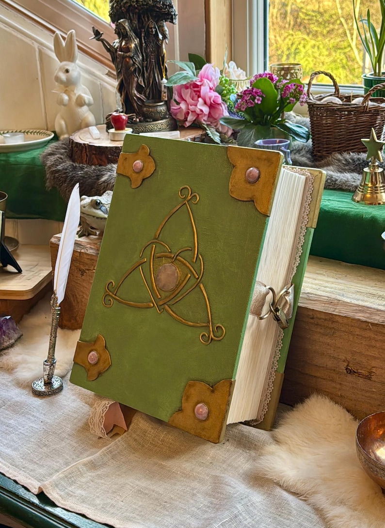 May include: A green book with a gold Celtic knot design and pink accents. The book has gold corner pieces and a ribbon closure. The pages are edged with lace. The book is propped open on a table with a white feather quill and other decorative items.