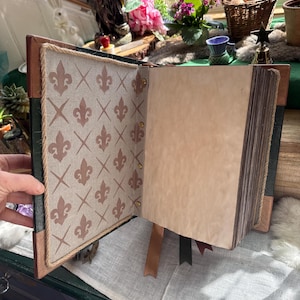 May include: Open journal with a brown leather cover and decorative interior pages. The left page features a beige fabric with a brown fleur-de-lis pattern. The right page is blank with aged paper. Brown and green ribbons mark pages.