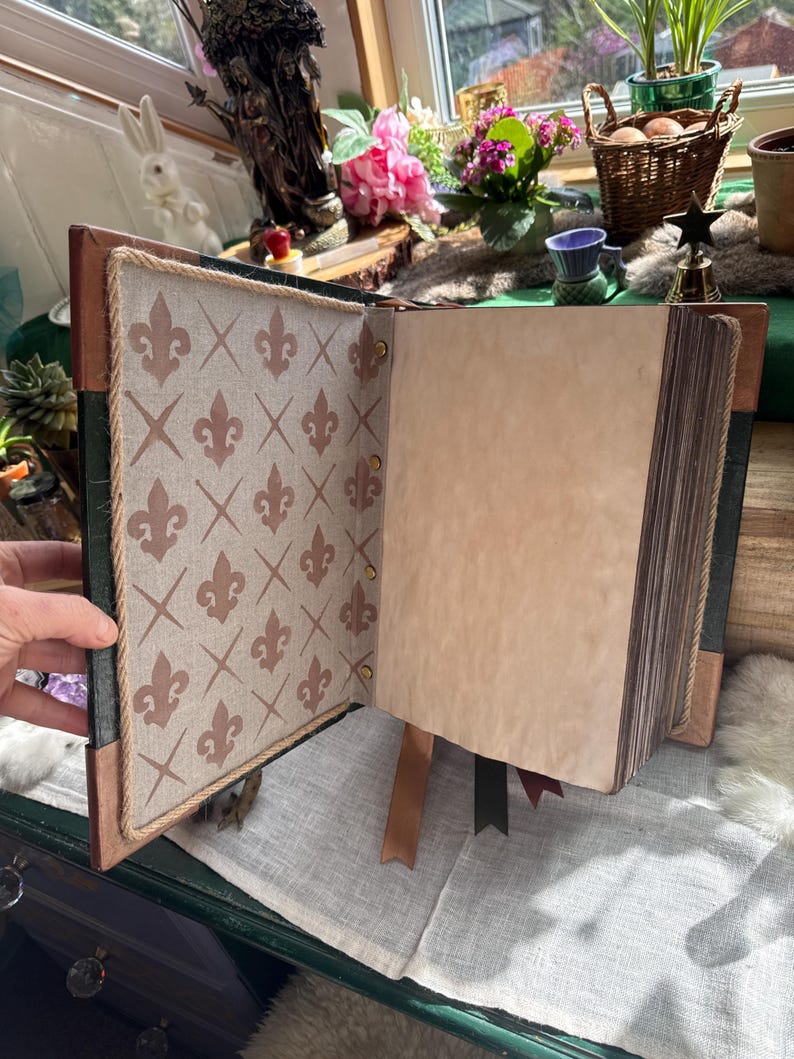 May include: Open journal with a brown leather cover and decorative interior pages. The left page features a beige fabric with a brown fleur-de-lis pattern. The right page is blank with aged paper. Brown and green ribbons mark pages.