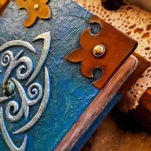 May include: Close-up of a handmade book with a textured blue cover and ornate silver embellishments. The book features brown leather corner accents with gold studs. The pages are visible, and the book is resting on a textured surface.