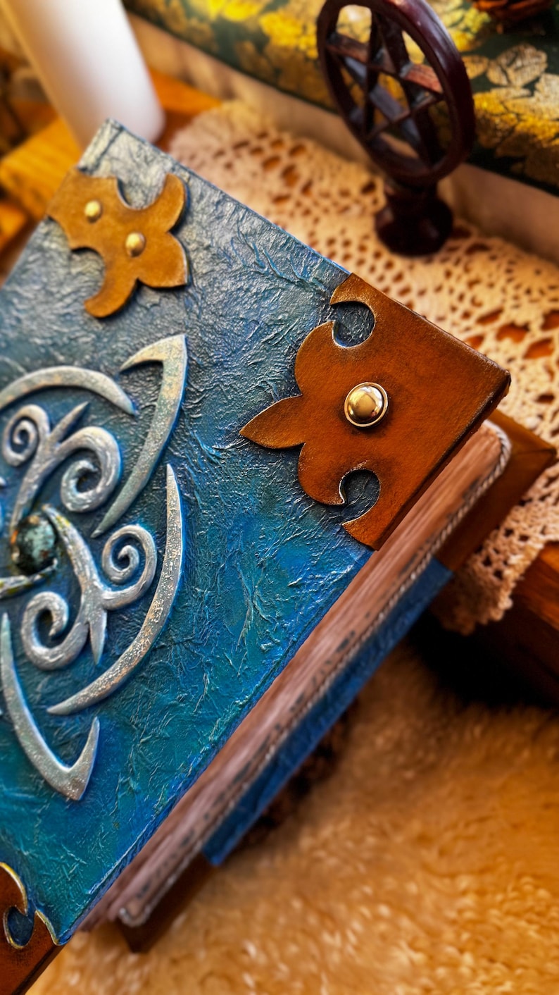 May include: Close-up of a handmade book with a textured blue cover and ornate silver embellishments. The book features brown leather corner accents with gold studs. The pages are visible, and the book is resting on a textured surface.
