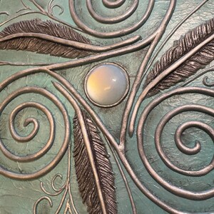 May include: Close-up of a teal-colored surface with silver-colored metal embellishments. The design includes swirling patterns, stylized feathers, and a central, oval-shaped, opalescent stone. The metalwork has a raised, three-dimensional effect.
