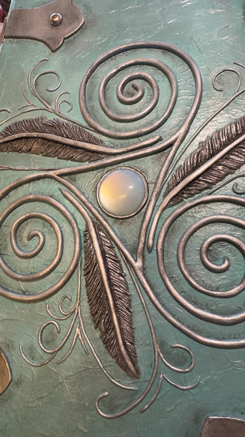 May include: Close-up of a teal-colored surface with silver-colored metal embellishments. The design includes swirling patterns, stylized feathers, and a central, oval-shaped, opalescent stone. The metalwork has a raised, three-dimensional effect.