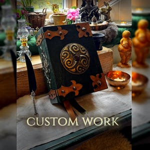May include: A handcrafted, dark green book with ornate gold and brown accents. The cover features a central circular design and decorative corner pieces. The text "CUSTOM WORK" is displayed at the bottom of the image. A quill pen rests nearby.