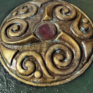 May include: Close-up of a handcrafted journal with a dark green cover. The cover features a bronze-colored, ornate circular emblem with a central red stone. Decorative bronze accents adorn the top.
