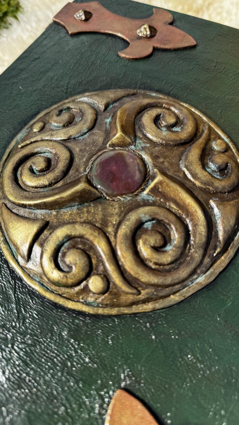 May include: Close-up of a handcrafted journal with a dark green cover. The cover features a bronze-colored, ornate circular emblem with a central red stone. Decorative bronze accents adorn the top.