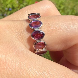 May include: A silver ring featuring a series of oval-cut, deep red gemstones. The ring is set in a silver band and is displayed against a blurred background of green foliage and colorful flowers. The gemstones have a rich, vibrant color.