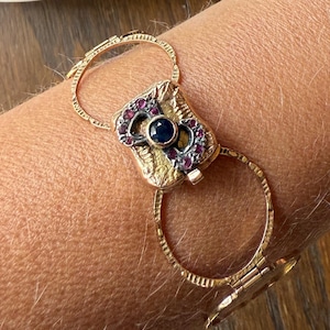 May include: Gold-toned bracelet with a central rectangular design featuring a dark blue gemstone and surrounding purple and clear stones. The bracelet is connected by two large circular links. A decorative plate with a floral design and a small wooden figurine are in the background.