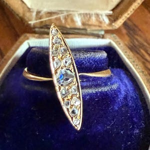 May include: A vintage gold ring with a marquise-shaped face, encrusted with small, clear and blue gemstones. The ring is displayed in an open, antique jewelry box with a blue velvet interior. The box is brown with gold lettering.