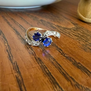 May include: A bypass ring featuring two round-cut blue sapphire stones set in a gold band, accented with small clear diamonds along the sides of the shank. The ring has a polished finish and a crossover design that holds the two primary blue stones.