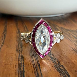 May include: A marquise-cut diamond ring with a ruby halo and gold band. The ring features a large, clear diamond center stone surrounded by rectangular rubies. The band has delicate detailing with small accent diamonds. The ring is set on a wooden surface.