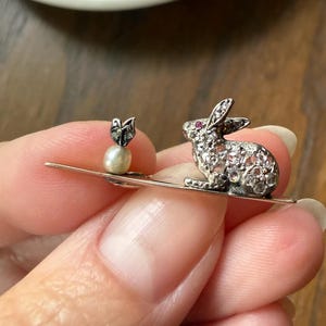 May include: A silver-toned bar brooch featuring a rabbit figure encrusted with clear stones and a small red eye, paired with a single white pearl topped by a small metallic floral element. The brooch is constructed on a thin, straight metal bar.