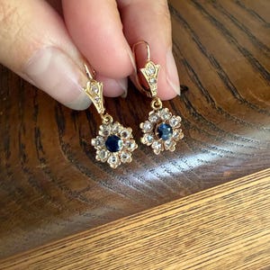 May include: A pair of gold drop earrings featuring a central round blue sapphire surrounded by a halo of small clear stones. The earrings have a lever-back closure with a small clear stone set into the top decorative metal component.