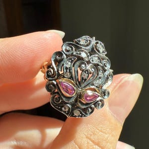 May include: An ornate ring featuring a central design with two pink gemstones and numerous small, clear stones. The ring has a silver-colored filigree setting with gold accents. The design is symmetrical and detailed, showcasing a vintage or antique style.