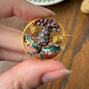 May include: A gold-toned brooch featuring a detailed thistle design. The brooch has a circular frame with a central thistle motif, accented with red and clear gemstones, and teal and gold enamel detailing. The brooch is held by a hand, with a decorative plate and small figurine in the background.