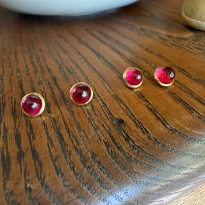 May include: Four round stud earrings featuring smooth, deep red cabochon stones set in simple gold-toned metal frames. The earrings are arranged in two pairs, each displaying a polished, domed surface that reflects light across the vibrant red color.