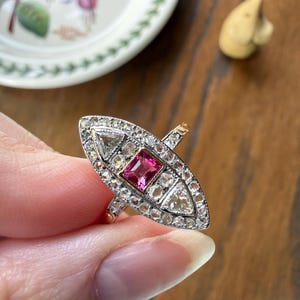 May include: An elongated marquise-shaped ring featuring a central rectangular pink gemstone flanked by two triangular diamonds, all surrounded by a border of small round diamonds set in a silver-toned metal frame on a thin gold-toned band.