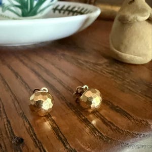 May include: A pair of gold-toned stud earrings featuring a spherical shape with a faceted, hammered texture. Each earring has a reflective surface that catches the light, creating multiple small highlights across the rounded, metallic exterior.