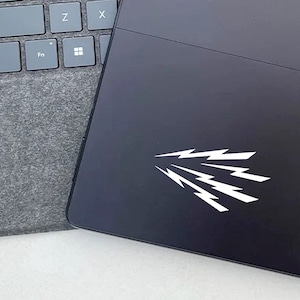 May include: A black tablet featuring a white graphic of multiple lightning bolts on its surface, positioned next to a gray fabric-covered keyboard with visible keys labeled Shift, Z, X, Ctrl, Fn, and a Windows logo icon.