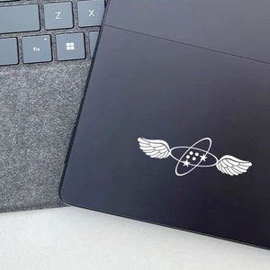 May include: A black laptop with a white sticker of a pair of wings surrounding a circle with three stars inside. The sticker is on the back of the laptop.