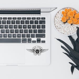 May include: A laptop with a black and white "AIR CREW" sticker featuring wings and a shield. The laptop is on a white surface, next to a small potted plant with orange flowers and a dark green plant.