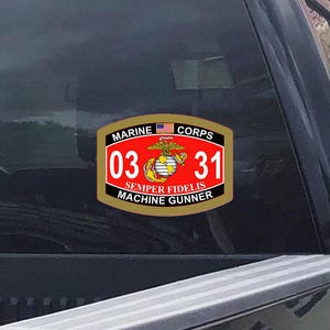 May include: A rectangular sticker with the words "MARINE CORPS" and a small American flag at the top. The center features the numbers "03" and "31" in white on a red background, with "SEMPER FIDELIS MACHINE GUNNER" below.