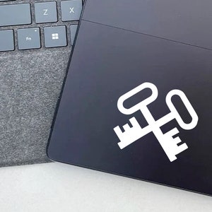 May include: A black laptop with a white crossed keys graphic. The laptop is partially open, revealing a gray keyboard and a dark gray felt cover. The keys graphic is a simple, bold design.
