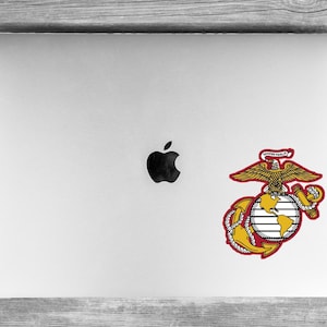 May include: A silver laptop with a black Apple logo in the center and a United States Marine Corps emblem sticker applied to the lower right corner. The emblem features a gold eagle, globe, and anchor with the text Semper Fidelis on a red banner.