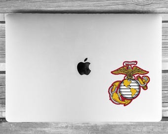May include: A silver laptop with a black Apple logo in the center and a United States Marine Corps emblem sticker applied to the lower right corner. The emblem features a gold eagle, globe, and anchor with the text Semper Fidelis on a red banner.