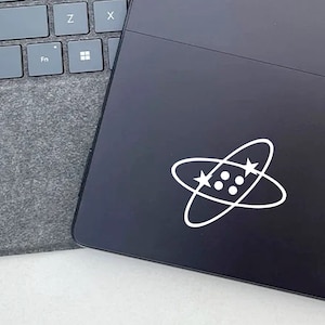 May include: A white sticker with a stylized atom design on a black laptop. The atom has four white dots in the center and two white stars on the outer rings.