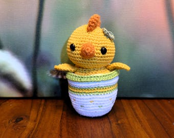 May include: A crocheted chick toy in a yellow and white egg-shaped container. The chick has a green flower on its head and black eyes.