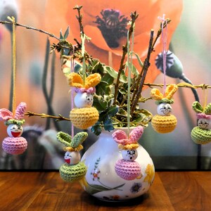 Op de afbeelding: Een witte vaas met bloemenmotieven bevat zes gehaakte konijnenversieringen. De konijnen zijn roze, geel en groen met lachende gezichten. De vaas is omringd door takken met groene bladeren.