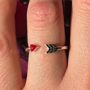 Puede incluir: Un anillo de plata con un corazón rojo y un diseño de flecha. El anillo se lleva en el dedo de una persona.