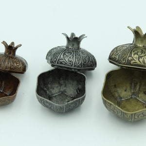 May include: Three ornate metal pomegranate-shaped boxes with lids open, revealing the interior. The boxes are in different sizes and finishes: copper, silver, and gold.