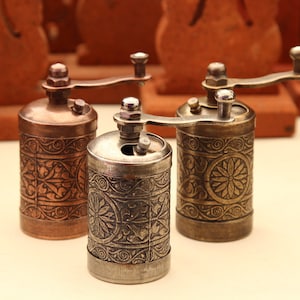 Puede incluir: Tres molinillos de pimienta de estilo antiguo con intrincados diseños florales. Los molinillos están hechos de metal y tienen un acabado plateado, cobre y latón.