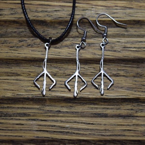 Op de afbeelding: Een zilverkleurige sieradenset met een hangerketting en een paar oorbellen. De hanger en oorbellen hebben een gestileerd, klauwachtig ontwerp. De ketting heeft een zwart koord. De sieraden worden gepresenteerd op een houten oppervlak.