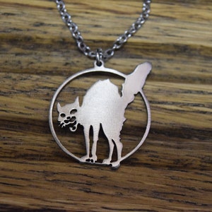 May include: A silver necklace with a circular pendant featuring a cutout silhouette of a black cat with its back arched and its tail raised. The cat is in a defensive posture, with its claws extended and its teeth bared.