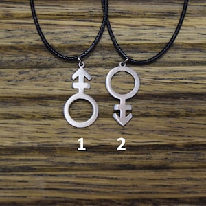 May include: Two silver necklaces with pendants on a wooden background. The pendants are the symbols for male and female. The necklaces are numbered 1 and 2.